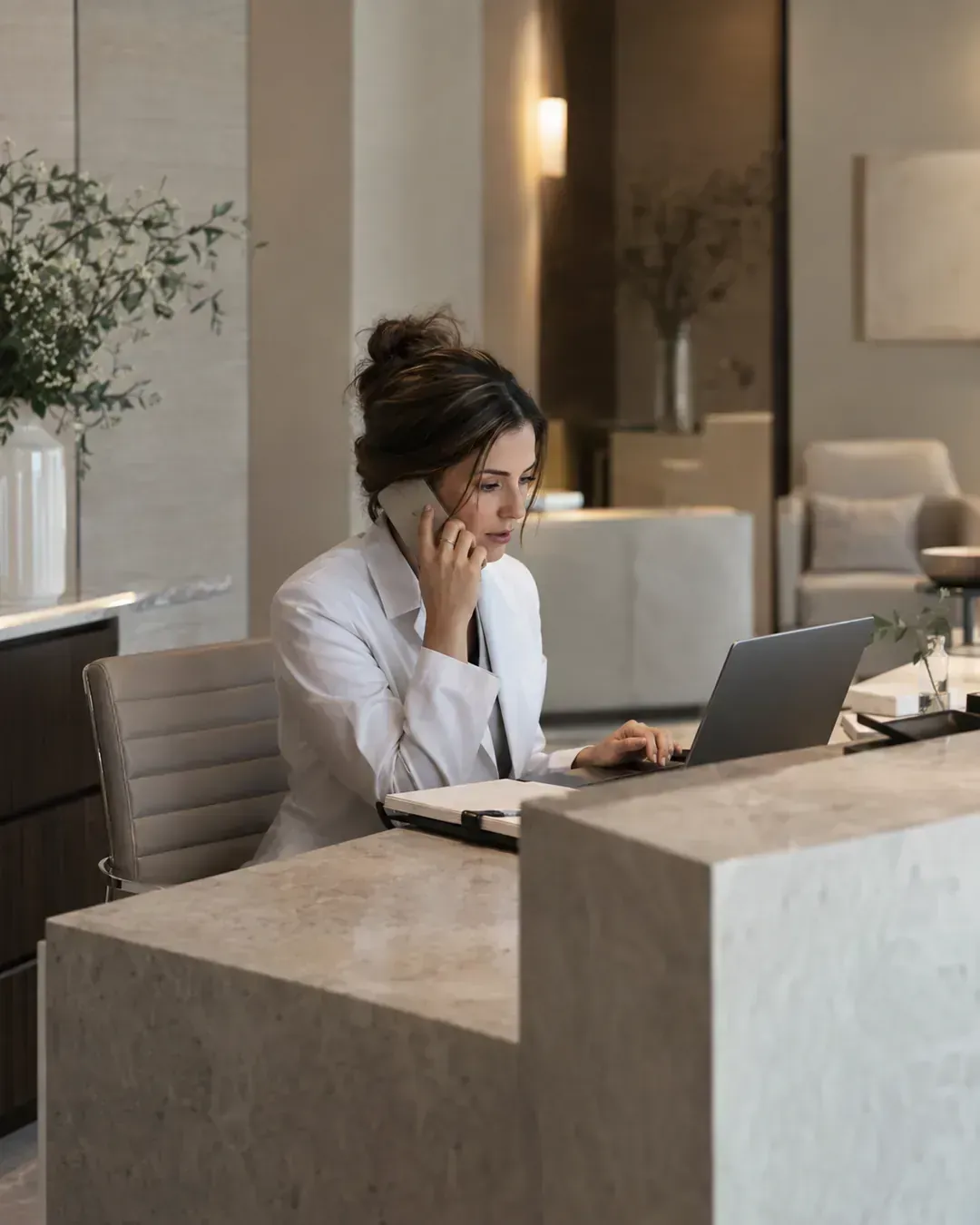 Recepcionista de clínica médica atendendo ao telefone e ao computador — atendimento de potenciais pacientes